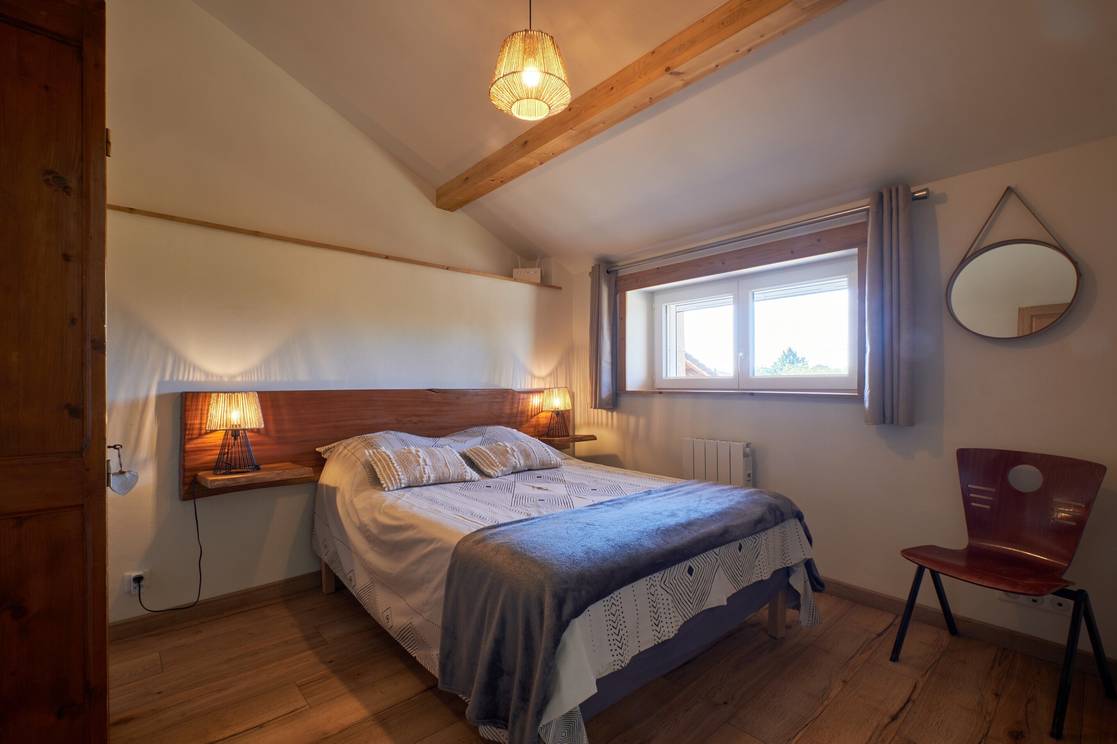 A bedroom with a bed, bedside lamps, a window with curtains, a chair, and a hanging mirror.