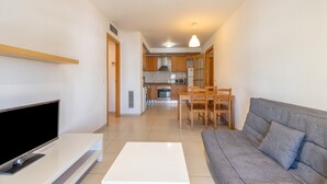 Ein modernes Wohnzimmer mit einem Flachbildfernseher, einem weißen Couchtisch und einem grauen Sofa. Im Hintergrund ist eine Essecke mit Holzkstühlen und einem Tisch erkennbar.