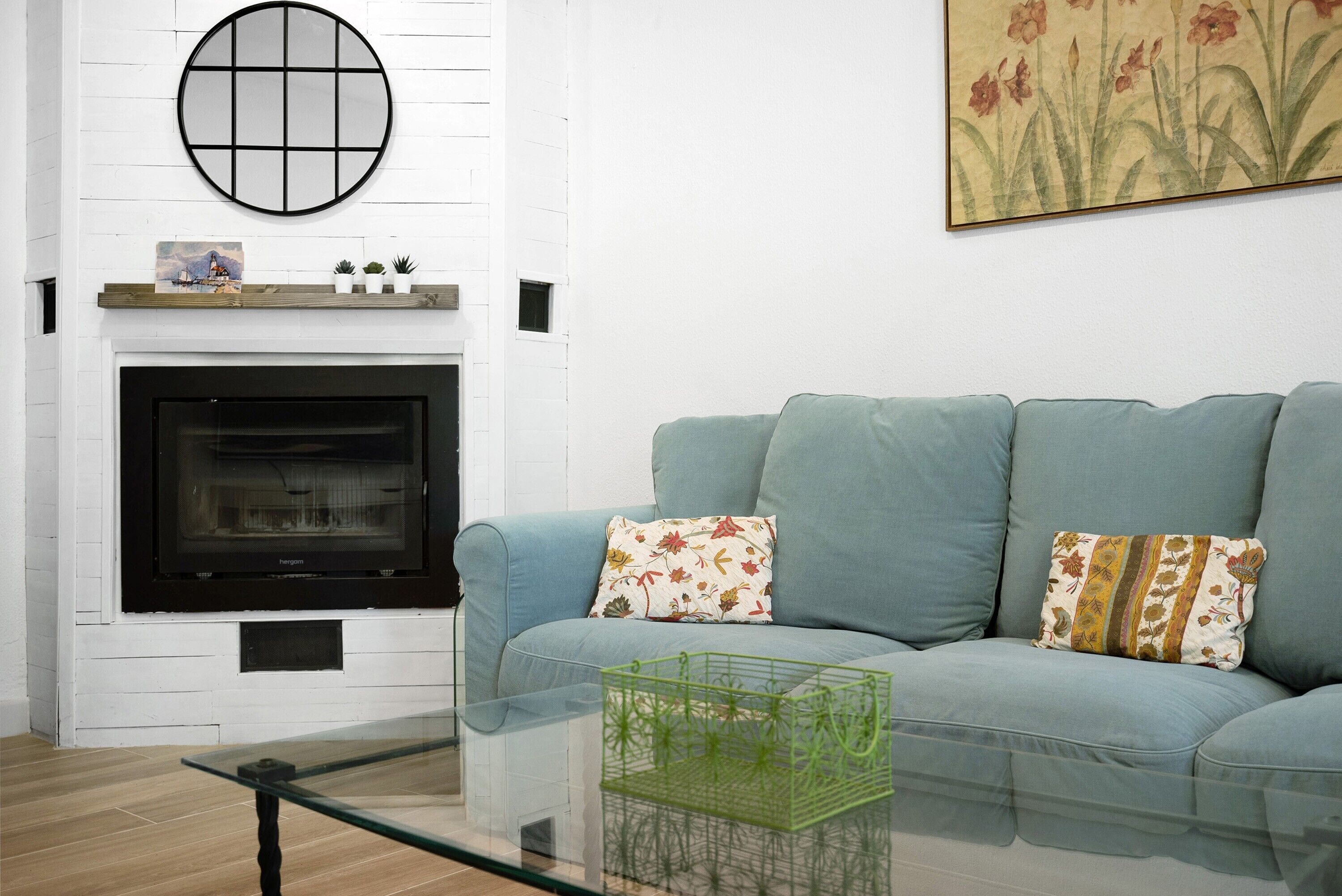 Una sala de estar con chimenea, un sofá con cojines estampados, una mesa de centro de vidrio y un contenedor verde.