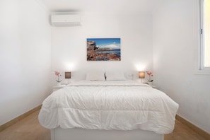A bedroom with a bed, a wall-mounted air conditioner, a painting of a coastal scene, two bedside lamps, and a nightstand with a flower.