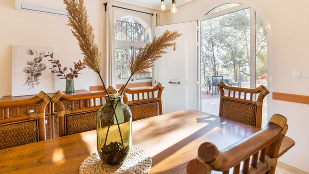 A dining area with a wooden table, chairs, a vase with dried plants, and a view through a glass door.