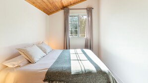 A bedroom with a wooden ceiling, a bed with white bedding, a window with curtains, and a hanging light fixture.