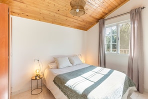 Ein Schlafzimmer mit Holzbalkendecke, einem Bett mit gestreifter Decke, einer Nachttischlampe und einem Fenster mit Vorhängen.