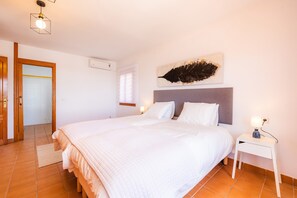 Een slaapkamer met een groot bed, een nachtkastje met een lamp, een kunstwerk aan de muur en een deur naar een andere kamer.