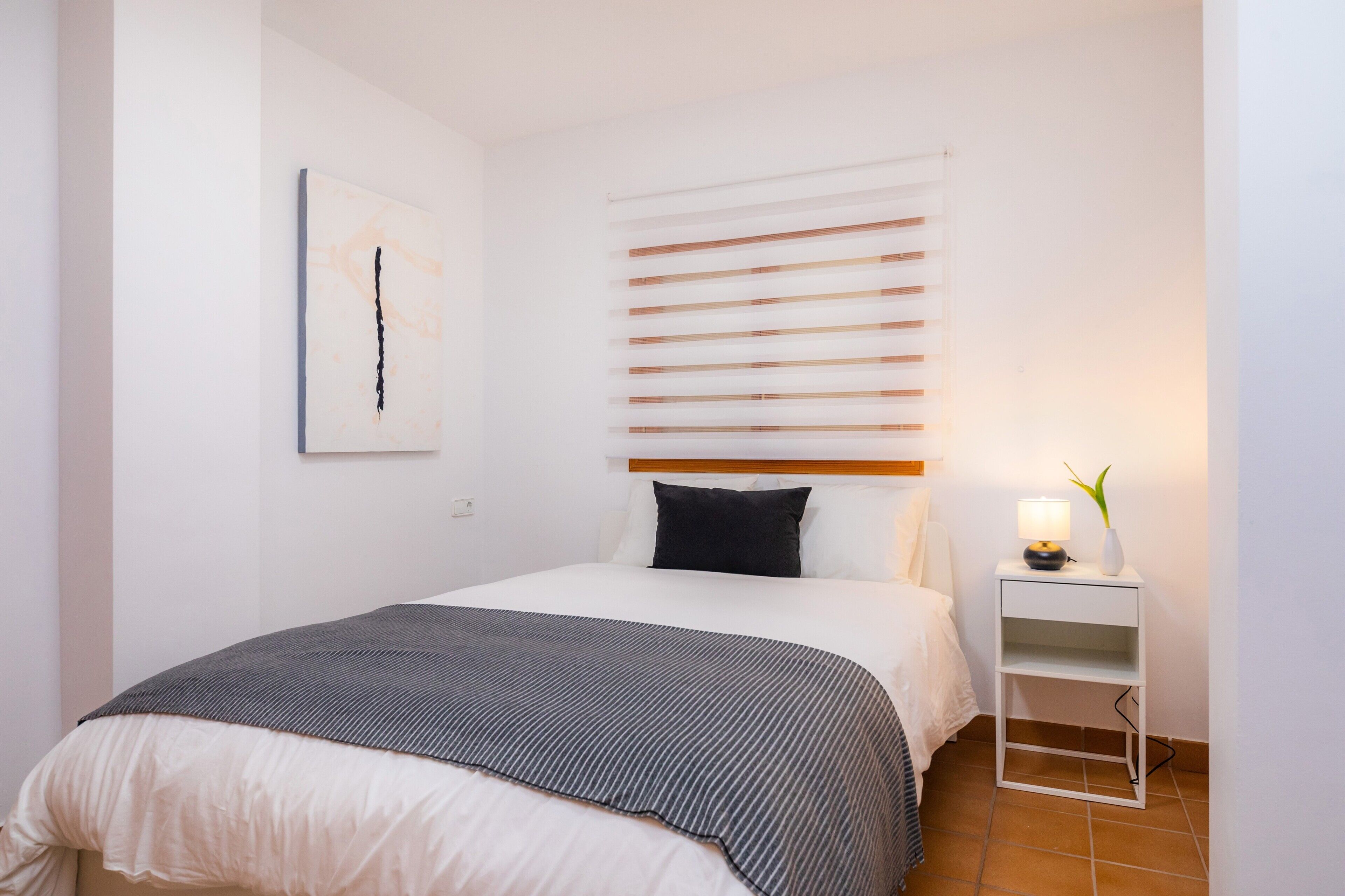 A neatly made bed with a white and gray striped blanket, a black pillow, and a white headboard. There is a bedside table with a lamp and a vase with a plant. A framed picture hangs on the wall.