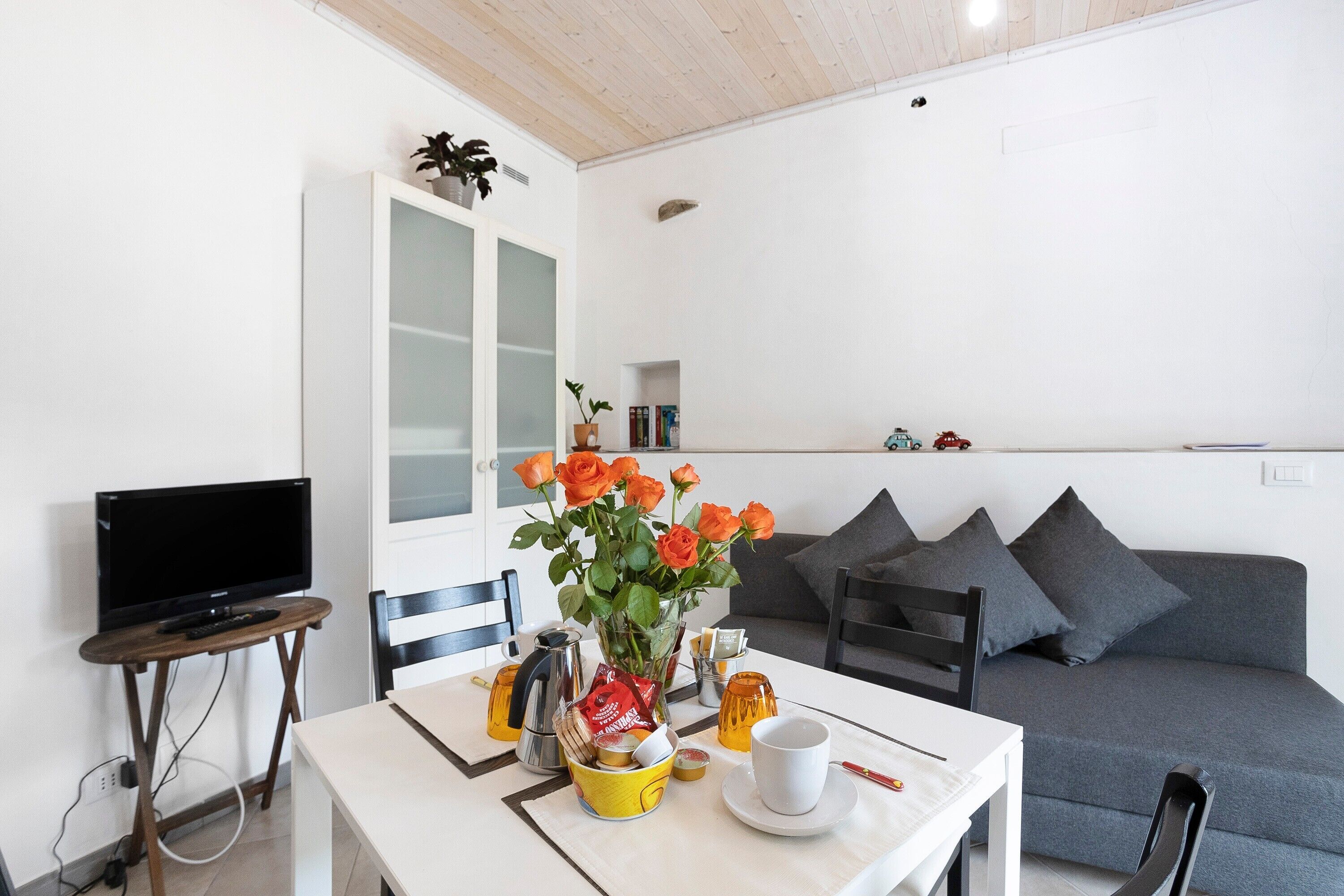 Uma sala de jantar com uma mesa, cadeiras, uma televisão, um sofá e um vaso com flores.