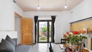 Ein Zimmer mit einer Sitzecke, einem Esstisch mit Stühlen, einer Küchenzeile und Blick ins Freie durch eine Tür.