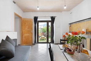 A room with a sofa, a dining table with chairs, a kitchen area, and a view through a door to the outside.