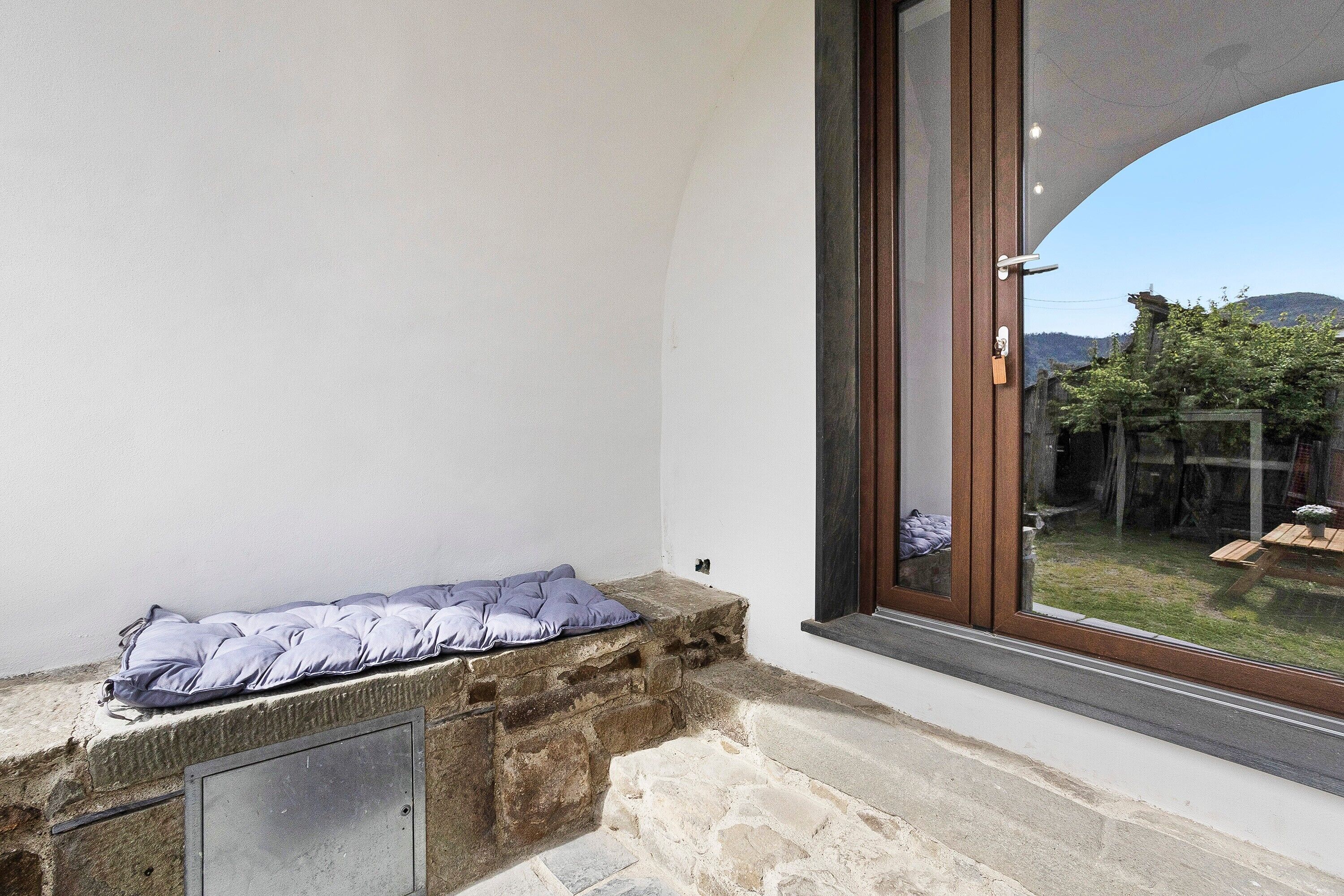 Un banco de piedra con cojín, una ventana con vistas a un jardín y una puerta con picaporte.