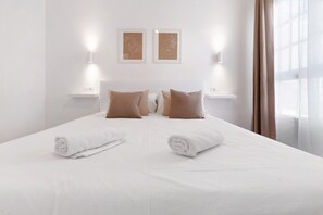 A neatly made bed with white linens and brown pillows, two wall-mounted light fixtures, and a window with sheer curtains.