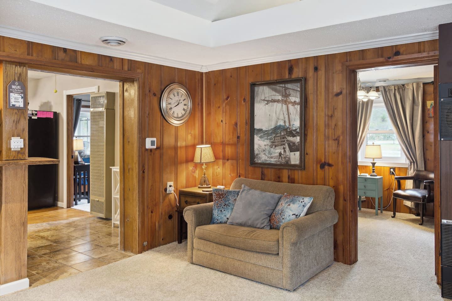 Une pièce chaleureuse avec un mur lambrissé en bois, une horloge, un tableau encadré, une lampe et un canapé avec des coussins.