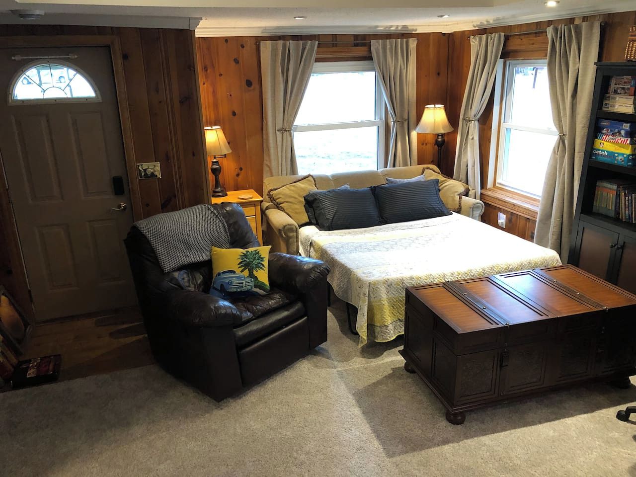 Una habitación acogedora con una cama, un sillón de cuero, un cofre de madera y una estantería.
