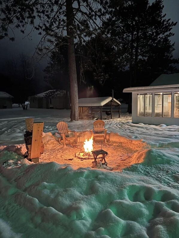 A snow-covered outdoor area with a fire pit, two chairs, and a wooden bench.