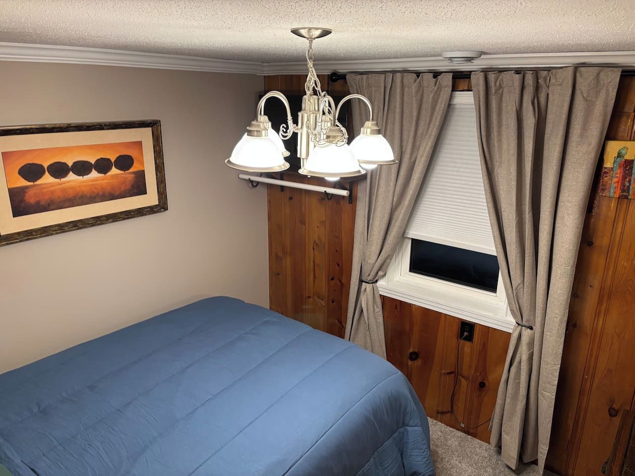Een slaapkamer met een bed, een raam met gordijnen, een wandlamp en een ingelijst kunstwerk.