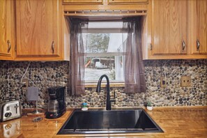 Una cocina con armarios de madera, salpicadero de piedra, ventana con cortina, cafetera y tostadora.