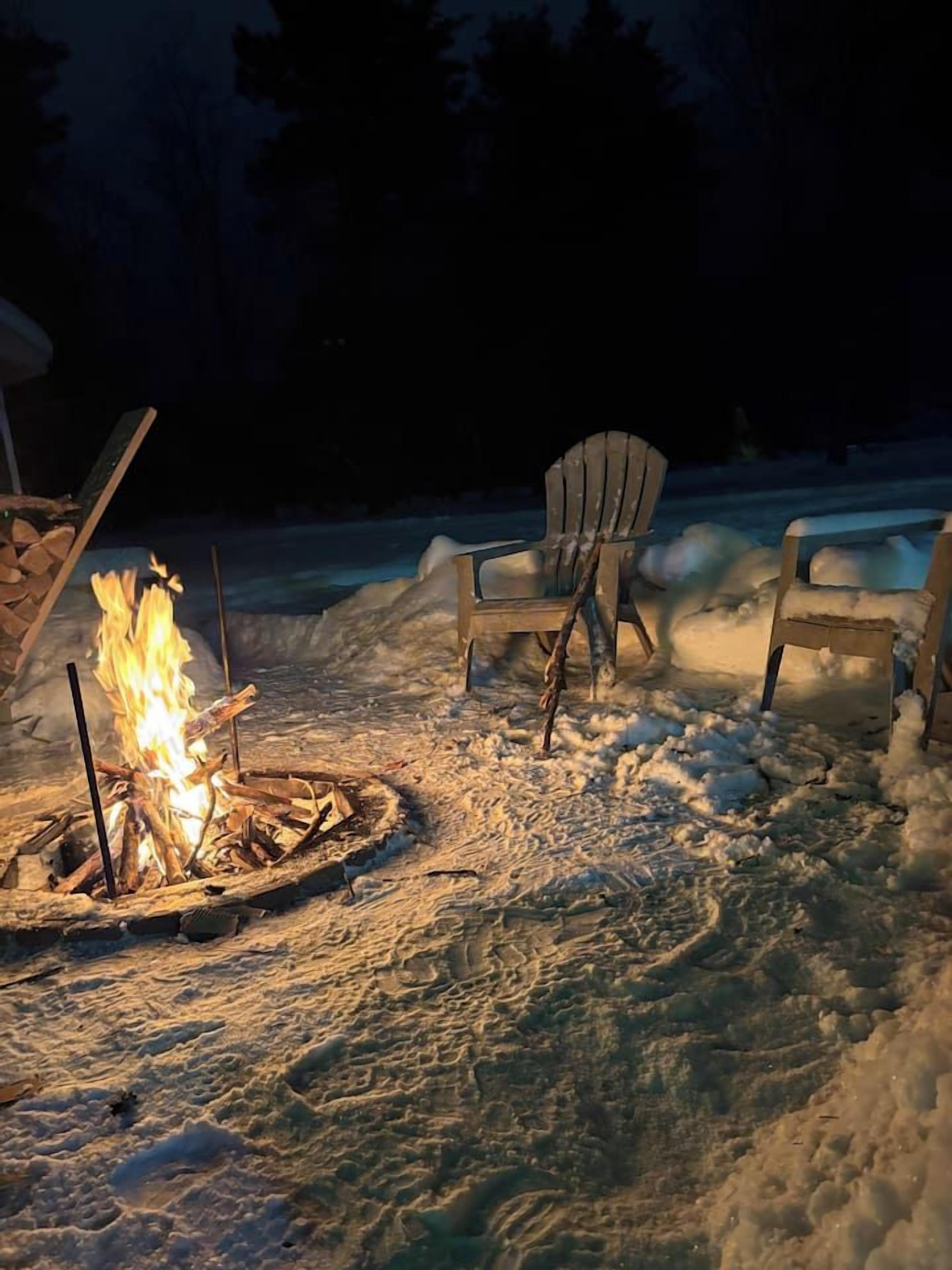 A snow-covered outdoor area with a campfire and two Adirondack chairs.