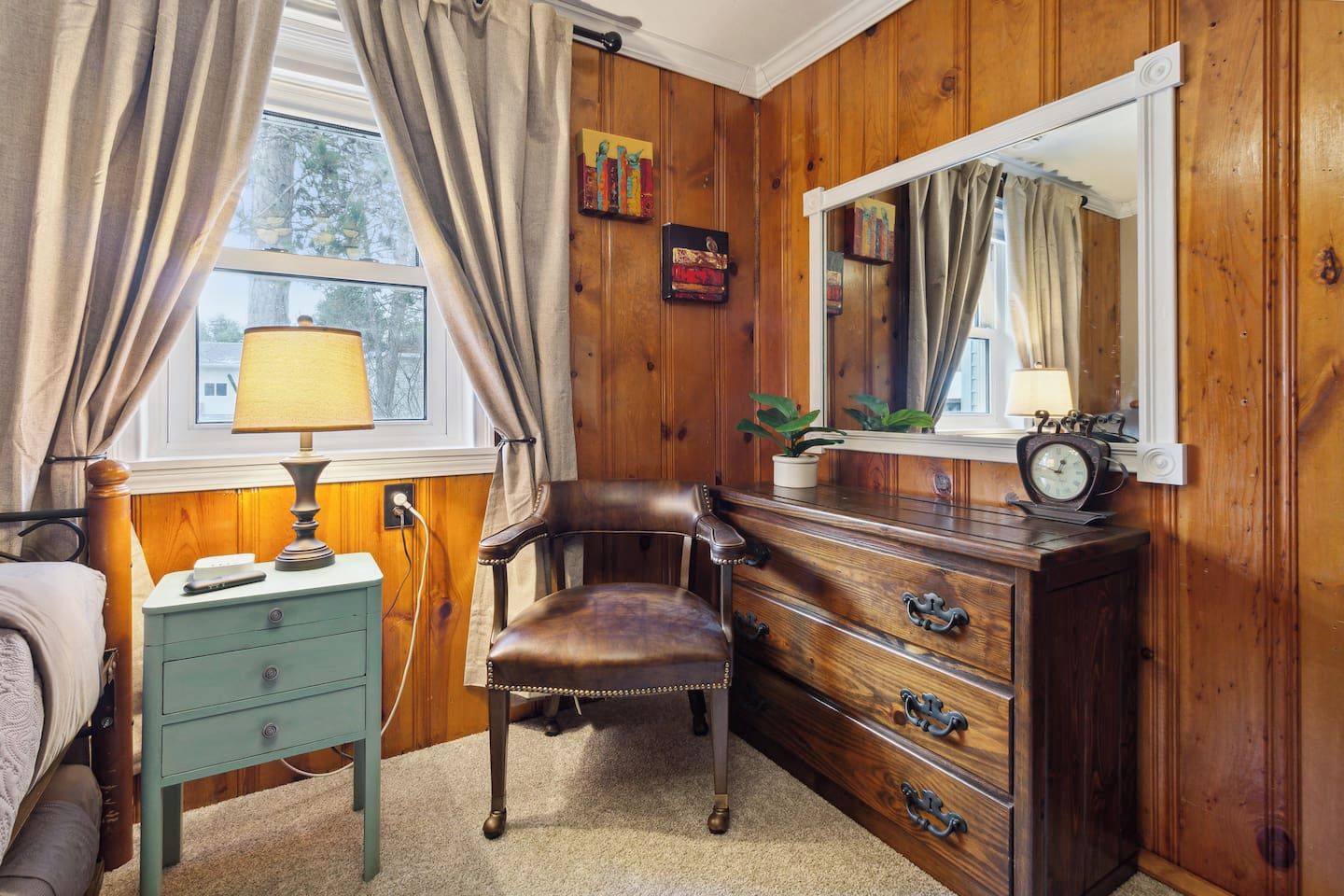 A room with wooden paneling, a bed, a dresser with drawers, a chair, a lamp, a mirror, and a clock.
