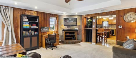 A spacious living room with a fireplace, wooden walls, a ceiling fan, and a kitchen visible through an open doorway.