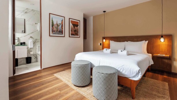 A modern hotel room with a wooden floor, a large bed with white linens, two ottomans, a wooden headboard, and framed pictures on the wall. - w* | Luminous 1BR with Amazing Deco in Polanco (Ciudad de México)