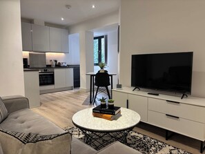 En moderne stue med sofa, sofabord med bøger, fjernsyn og et køkken i baggrunden.