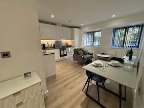 Une cuisine moderne avec des meubles blancs, un coin repas avec une table blanche et des chaises noires, et une vue sur l’extérieur par une fenêtre.