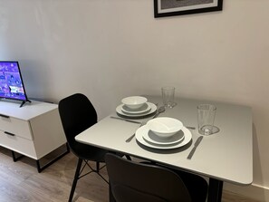 Une table à manger dressée avec de l’assiette, des bols et des couverts blancs, une chaise noire et un tableau encadré accroché au mur.