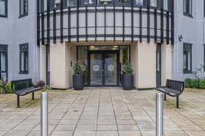 Une entrée d’immeuble avec une façade incurvée, deux plantes en pot et deux bancs au premier plan.