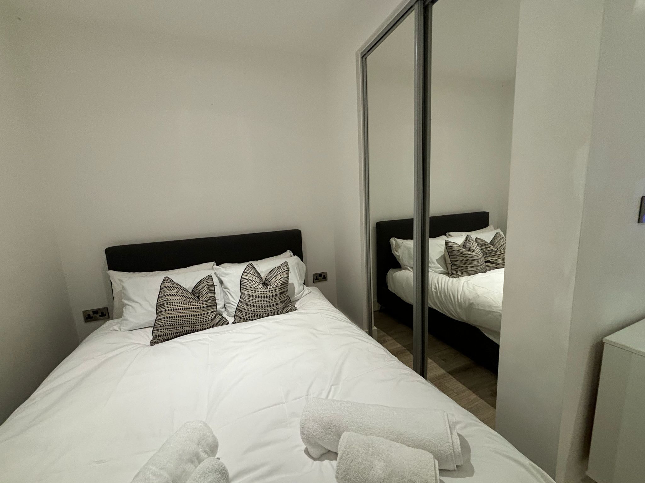 A hotel room with a single bed, white bedding, a black headboard, and a mirror reflecting the room.