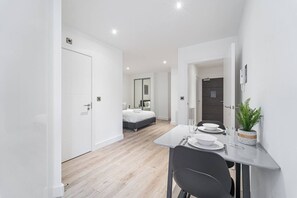 Een moderne, minimalistisch ingerichte kamer met een bed, een eethoek met een tafel en stoelen, en een kleine plant in een pot.