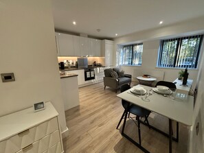 Une cuisine moderne avec des meubles blancs, un coin repas avec une table blanche et des chaises noires, et une vue sur l’extérieur par une fenêtre.
