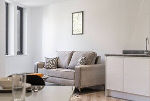 Un salon moderne avec un canapé gris, une table à manger et un coin cuisine doté de meubles blancs et d’un évier.