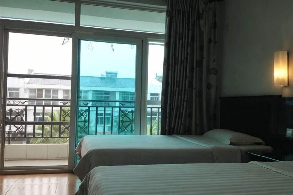 A hotel room with two beds, a balcony with a view of buildings, and a nightstand with a lamp.