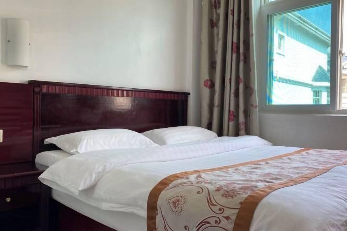 A hotel room with a wooden headboard, a large window with curtains, and a bed with white bedding and a decorative throw.