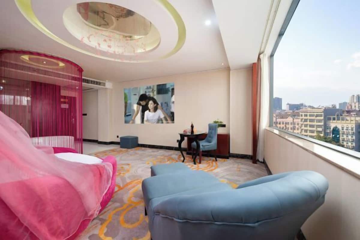 Habitación de hotel con vista a la ciudad, una cama con dosel rosa, dos sillas azules, una mesita y una lámpara en el techo.