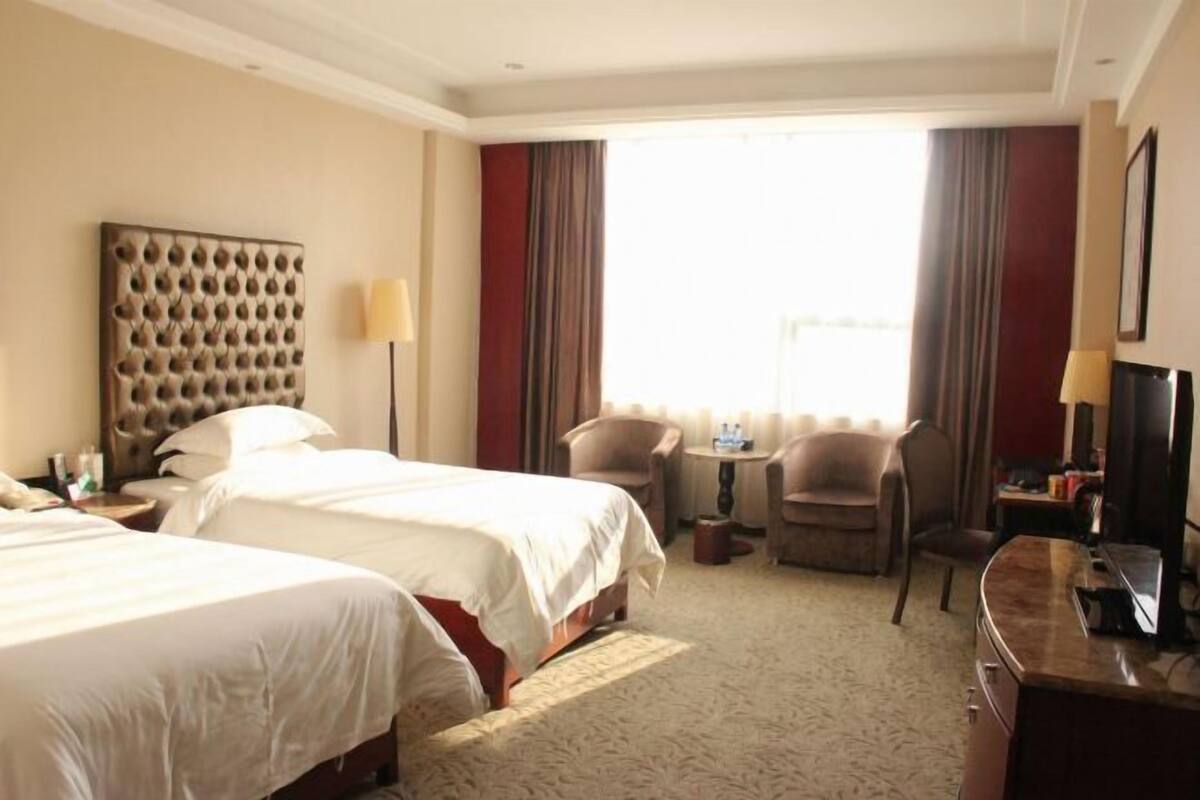Habitación de hotel con dos camas, un cabecero grande y capitonado, una pequeña mesa con sillas, un escritorio con televisor y un amplio ventanal con cortinas.
