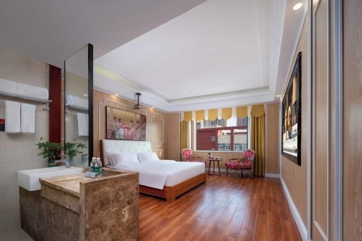 Habitación de hotel con una cama grande, zona de estar con una silla y una mesa, baño con lavamanos y espejo, y vistas de edificios a través de la ventana.