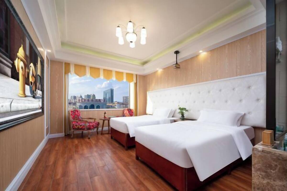 Habitación de hotel con dos camas, un ventanal amplio con vista a la ciudad, una araña y suelo de madera.