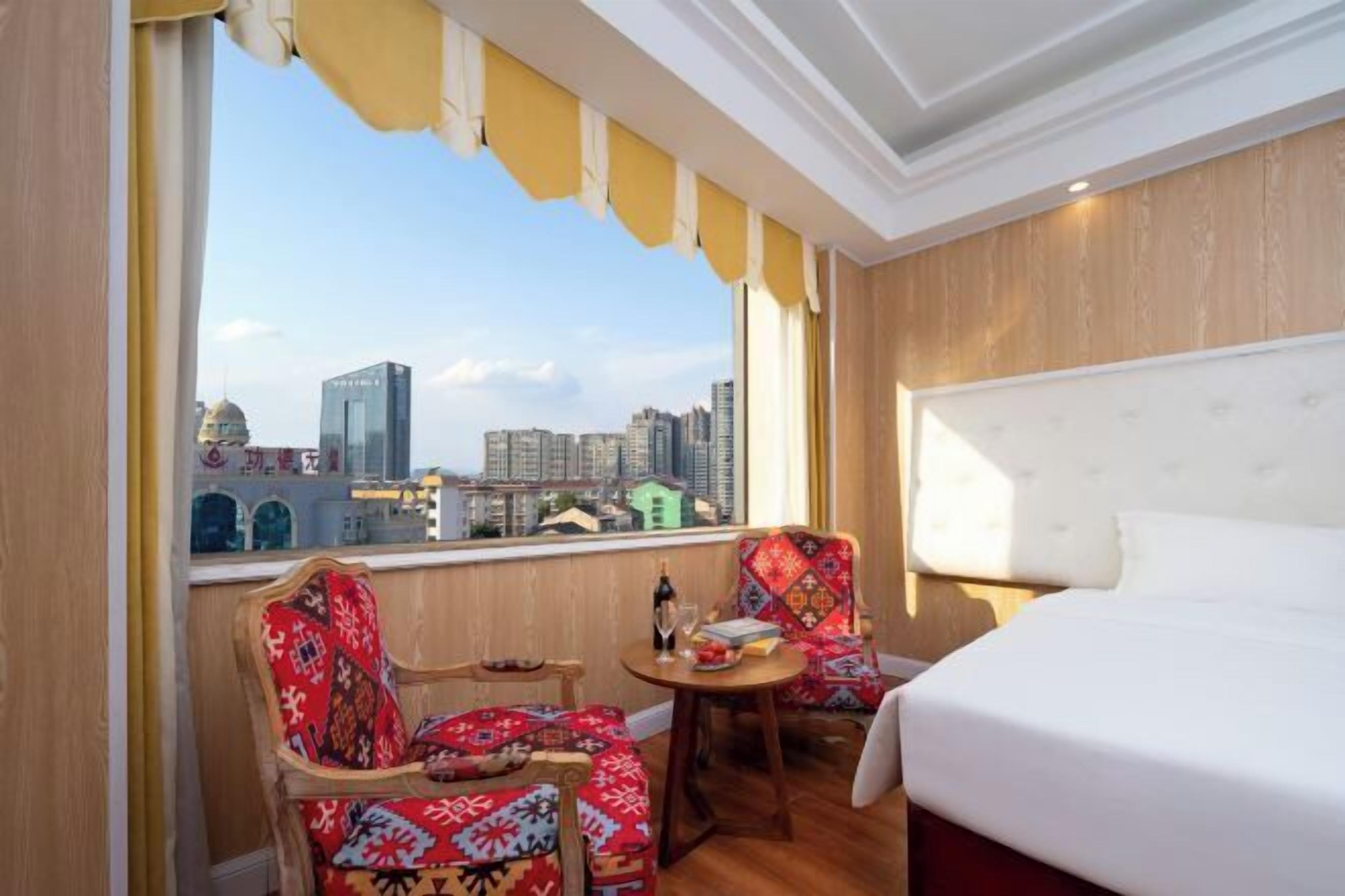 A hotel room with a large bed, two chairs, a small table, and a view of the cityscape.