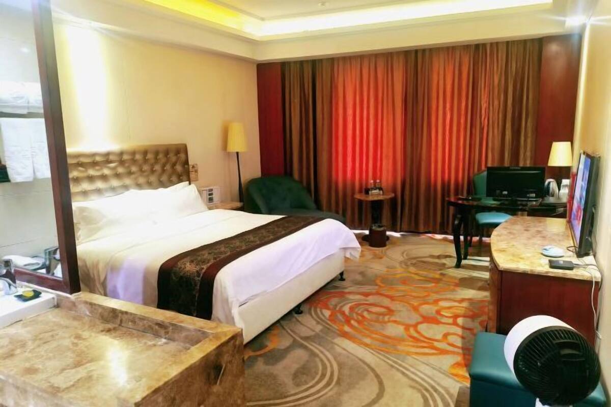 Habitación de hotel con cama, escritorio, silla, mesita, ventilador y ventana grande con cortinas.