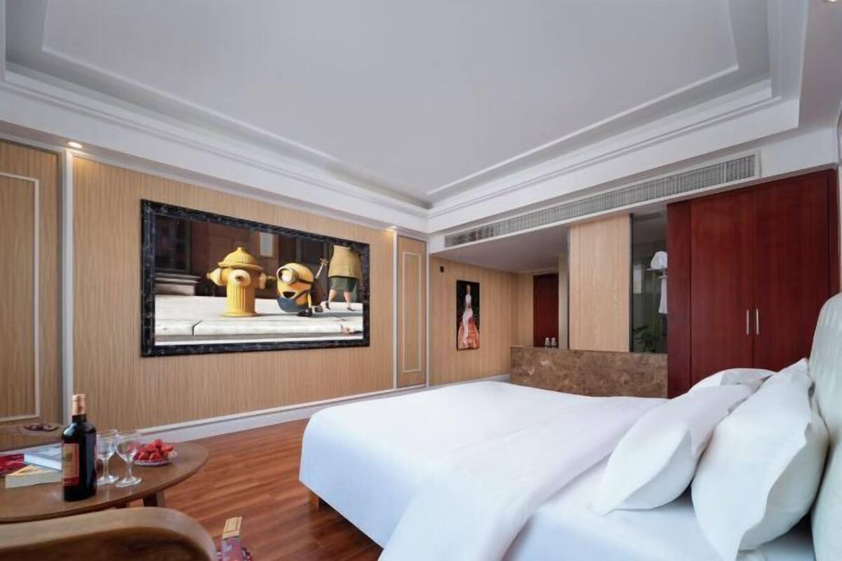 Habitación de hotel con una cama grande, una mesa redonda con una botella y vasos, un cuadro en la pared y un espejo que refleja la habitación.