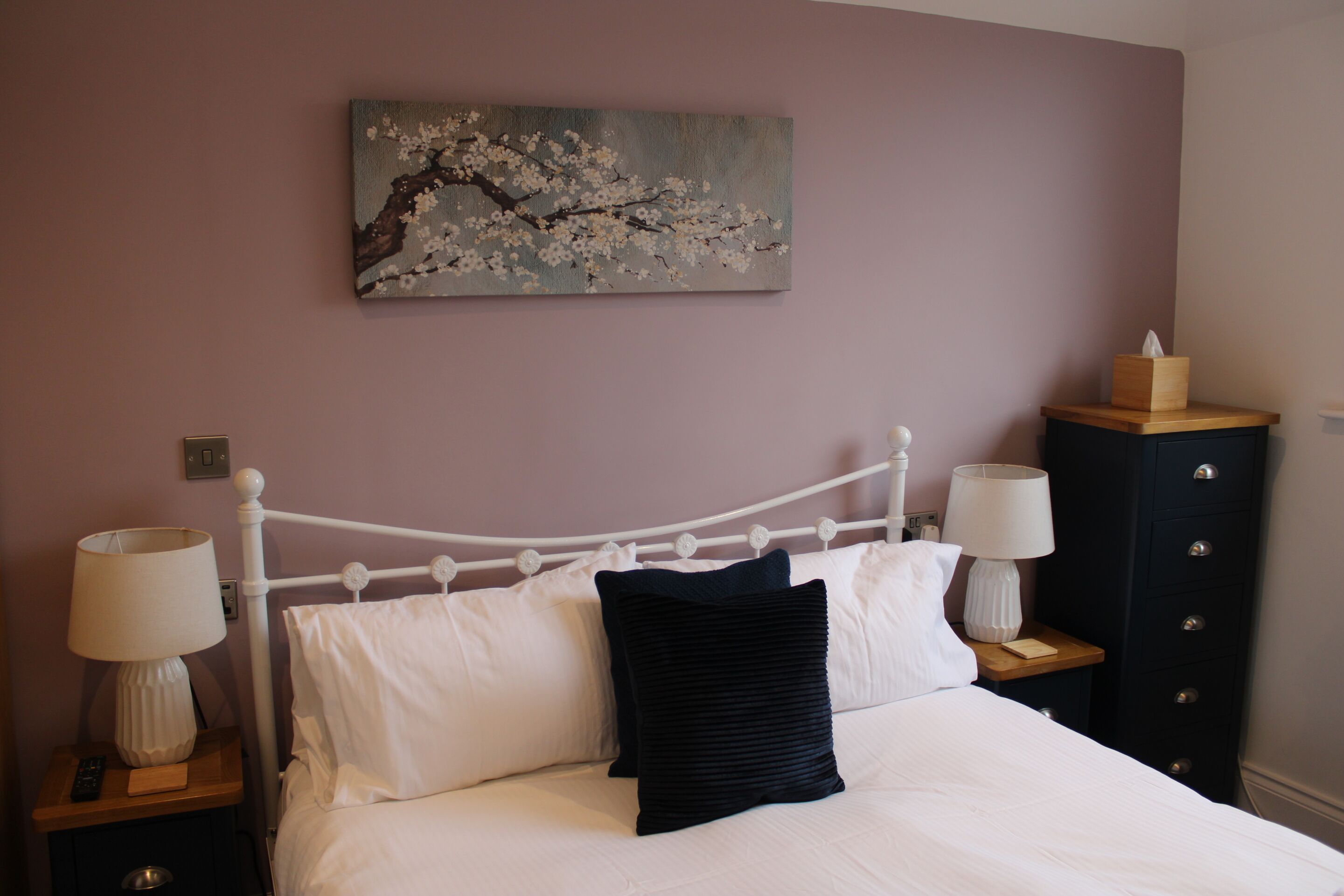 A neatly made bed with white linens and a black pillow, flanked by two bedside tables with lamps and a wooden nightstand with a tissue box. A painting of a tree branch with blossoms hangs above the bed.