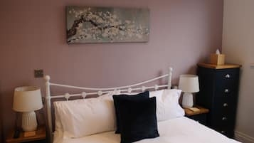 A neatly made bed with white linens and a black pillow, flanked by two bedside tables with lamps and a wooden nightstand with a tissue box. A painting of a tree branch with blossoms hangs above the bed.