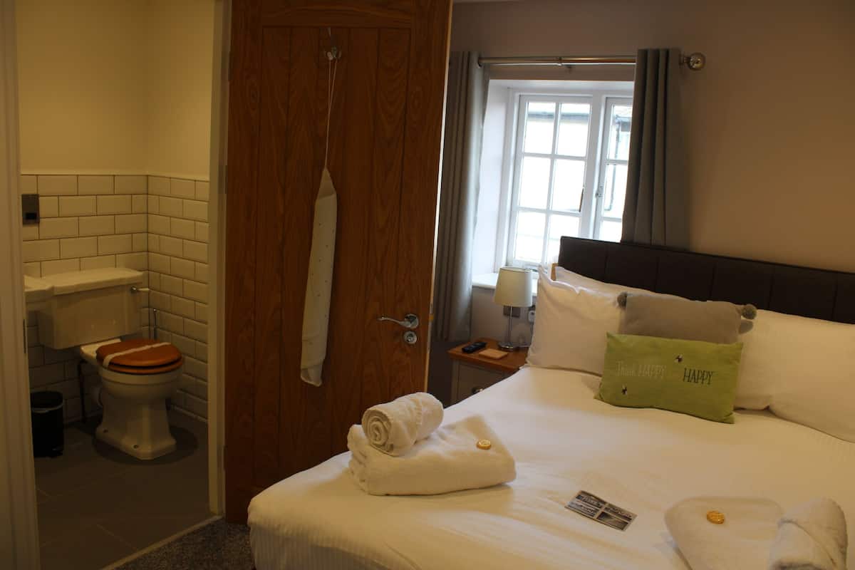 Habitación de hotel con puerta de madera, inodoro blanco, cama con ropa blanca, almohada verde y ventana con cortinas.