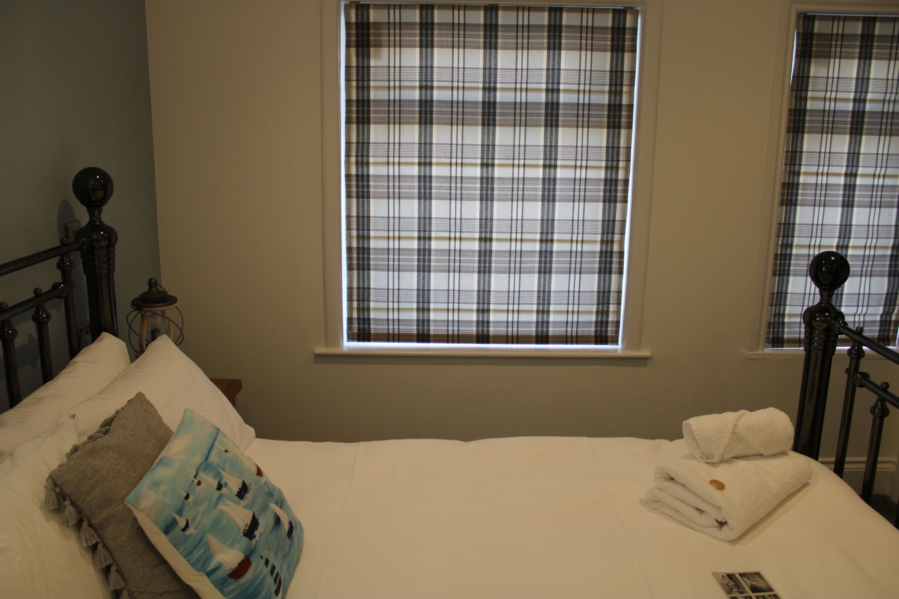 Una cama bien hecha con ropa de cama blanca, una almohada azul con diseño náutico y una toalla blanca doblada. Hay una ventana con cortina a cuadros y una mesita de noche con una lámpara junto a la cama.