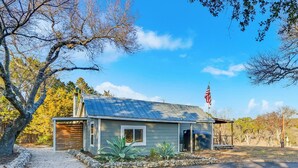 A single-story house with a metal roof, a flagpole with an American flag, and a stone pathway leading to the entrance. - Bungalow on the Bluff | Hot tub, Pet Friendly! (Wimberley)