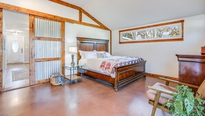 A bedroom with a wooden bed, a nightstand, a chair, and a large window. - Bungalow on the Bluff | Hot tub, Pet Friendly! (Wimberley)