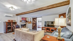 A spacious living room with a fireplace, a large sofa, a television, and a wooden ceiling beam. - Bungalow on the Bluff | Hot tub, Pet Friendly! (Wimberley)