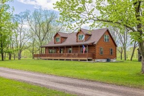 A wooden cabin with a porch, surrounded by trees and a grassy area.