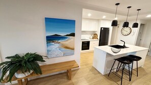 A modern kitchen with white cabinets, a central island, pendant lights, and a clock on the wall. - Jubilee Apartment 05 - Bring your own linen (South West Rocks)