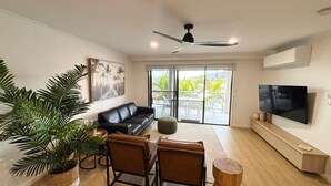 A living room with a black leather sofa, a television, a ceiling fan, and a large window with a view of palm trees. - Jubilee Apartment 05 (South West Rocks)